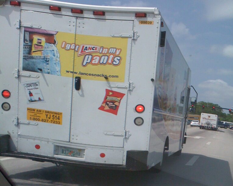 A white delivery truck featuring promotional advertisements for Lance Snacks boldly displays the slogan, "Lance in your Pants? Really? Is that Needed to sell Snacks?" The rear of the truck also sports a "How am I driving?" sticker and a license plate.