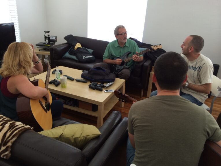 Four people sit on couches in a living room, playing and discussing guitars. Various electronics and bags are on the coffee table and couch, as they share insights from their Guitar Lessons at PaperSteet.