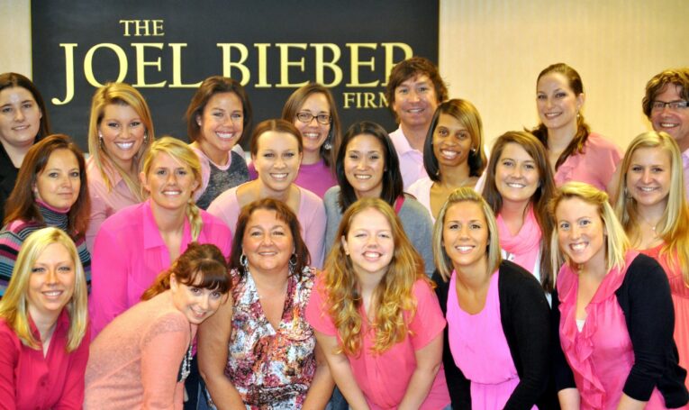 A group photo shows 20 people posing together, mostly smiling, in front of a sign that reads "The Joel Bieber Firm." Many are wearing pink tops.