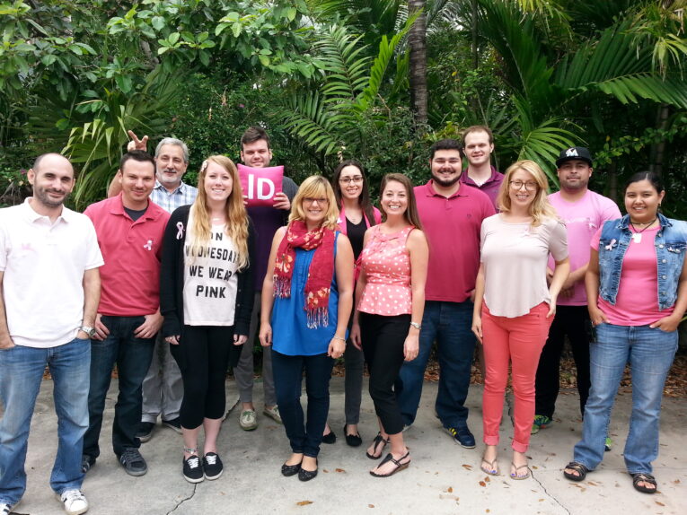 A group of thirteen people is standing outdoors in front of green foliage, several of whom are wearing pink clothing.