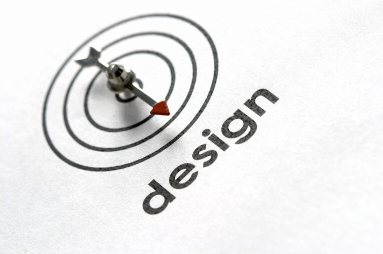 A dart with a red tip is embedded in a target graphic on a white paper, symbolizing precision. The word "design" is printed next to it, emphasizing the focus on web design. This visual metaphorically represents hitting all points on your comprehensive web design launch process checklist.