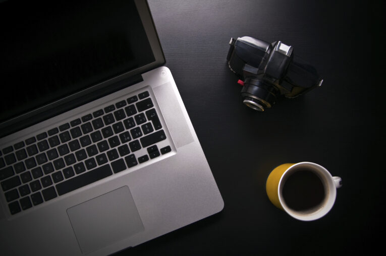 A laptop, a camera, and a cup of black coffee on a dark surface.