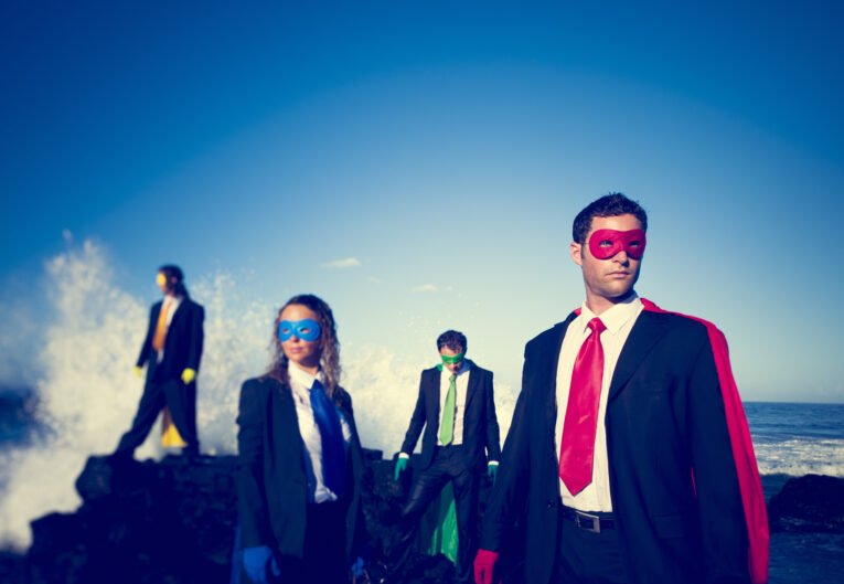 Four adults in suits and superhero masks stand on rocks by the ocean, with waves crashing behind them under a clear blue sky.