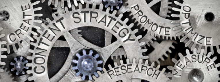 Close-up image of interlocking gears with words like "Content Strategy," "Create," "Research," "Promote," "Optimize," and "Measure" engraved on them, illustrating interconnected marketing concepts.