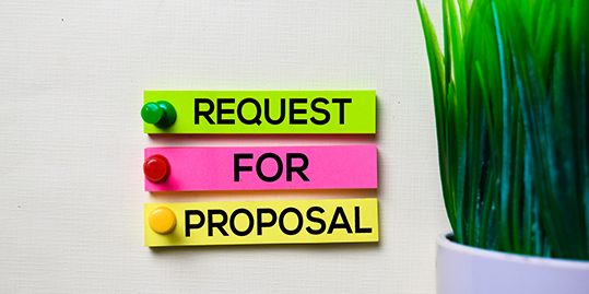 Three colorful strips with the words "REQUEST FOR PROPOSAL" pinned on a wall next to a green plant.