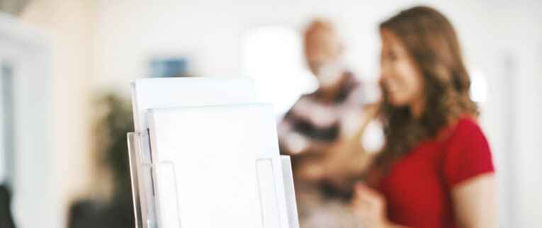 A display stand with white brochures in the foreground, with two out-of-focus individuals standing and talking in the background.