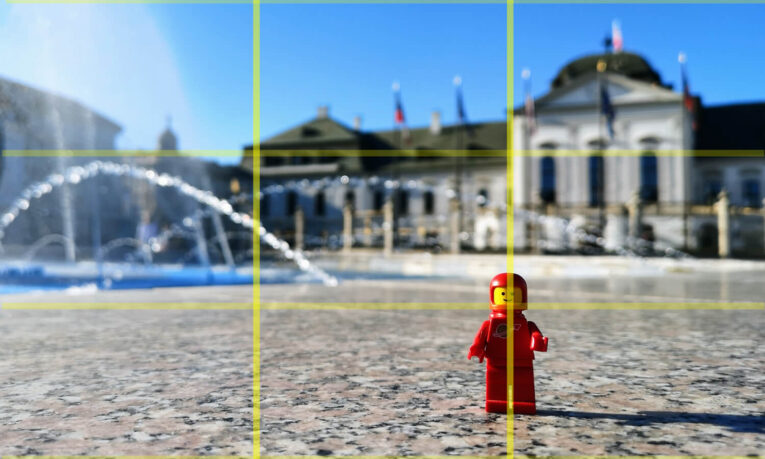 A red Lego astronaut figure stands on a granite surface in front of an ornate building with fountains and flags, captured in bright daylight.