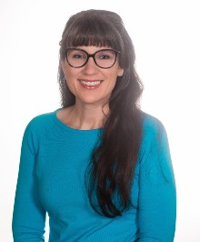 Woman with long dark hair, wearing glasses and a blue sweater, smiling in front of a white background as PaperStreet celebrates International Women's Day with our clients.