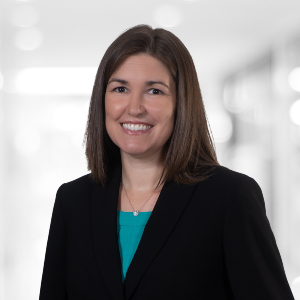 A woman with shoulder-length brown hair is smiling at the camera. She is wearing a black blazer over a teal top. The background, reflecting the festive spirit of "PaperStreet Celebrates International Women's Day with Our Clients," is blurred and brightly lit.