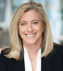 A smiling woman with long blonde hair wearing a white blouse and dark blazer, posing indoors with a blurred background, as PaperStreet celebrates International Women's Day with our clients.