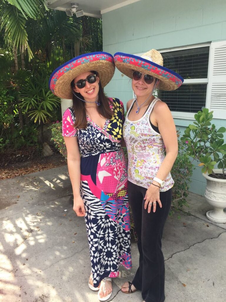 Two women wearing large sombreros and sunglasses stand side by side in a sunny outdoor setting, one in a colorful dress and the other in a printed tank top and black pants.